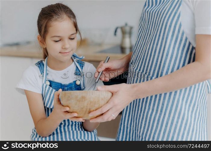Busy little girl hold bowl, looks how mother whisks ingredients, learns how to cook. Unrecognizable mother in striped apron spends time with daughter, shows how to prepares dough. Cooking time