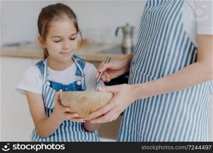 Busy little girl hold bowl, looks how mother whisks ingredients, learns how to cook. Unrecognizable mother in striped apron spends time with daughter, shows how to prepares dough. Cooking time