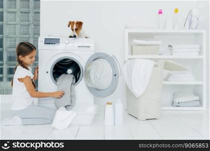 Busy little pretty girl does laundry at home, poses on knees near washing machine, uses liquid powder, pedigree dog on top of washer, looks at small female host householder. Housekeeping concept
