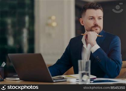 Busy male entrepreneur in dark blue suit sitting at his work desk in stylish office and thinking about new solutions to business challenges, holding pencil while being serious and concentrated. Thinking businessman sitting at his desk in front of laptop holding pencil