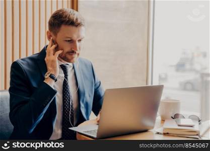 Busy male finance accountant with earbuds dressed in blue suit sitting alone in cafe, working through laptop, helping and explaining everything to his client in online voice meeting. Male finance accountant working online through laptop