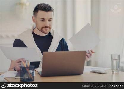 Busy male freelancer in casual clothes sitting at his workplace with computer and doing paperwork, analyzing project results while working remotely from home. Freelance and business concept. Focused businessman working with documents and using laptop
