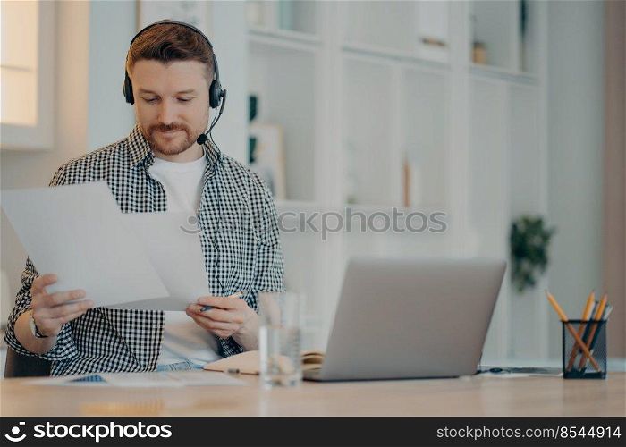 Busy male freelancer wearing headset holding documents in hands and working remotely on laptop while sitting in living room at home. Business online and freelance concept. Smiling young man working with documents at home