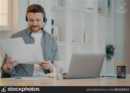 Busy male freelancer wearing headset holding documents in hands and working remotely on laptop while sitting in living room at home. Business online and freelance concept. Smiling young man working with documents at home