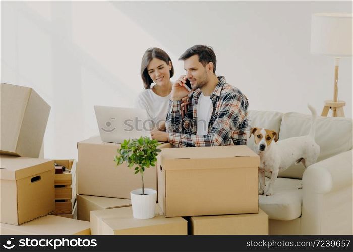 Busy male homeowner calls in delivery service via modern smartphone, makes order online, focused in laptop, caring wife helps husband to solve domestic problems, dog poses near on sofa, boxes around