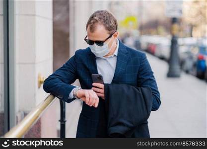 Busy man waits for business partner, looks at watch poses at street holds modern cellular, waits for call, wears medical mask, protects from coronavirus, cares about health during epidemic or pandemic