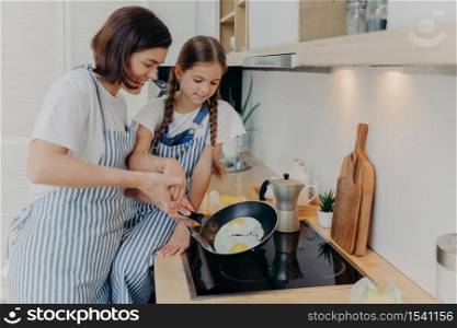 Busy mother and daughter wear striped aprons, pose at kitchen near cooker, fry eggs on pan, prepare fast breakfast, enjoy domestic atmosphere. Mum teaches small kid to cook. Happy family concept