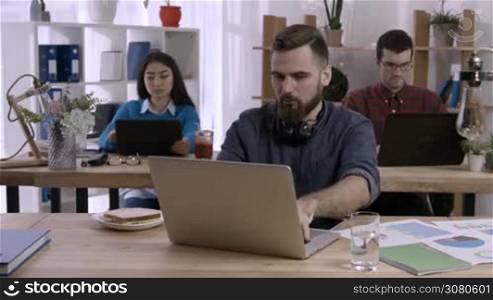Busy office worker eating a sandwich at his desk while working on laptop during lunch break hour in office. Young bearded hipster having a sandwich at workplace as his colleagues working on background