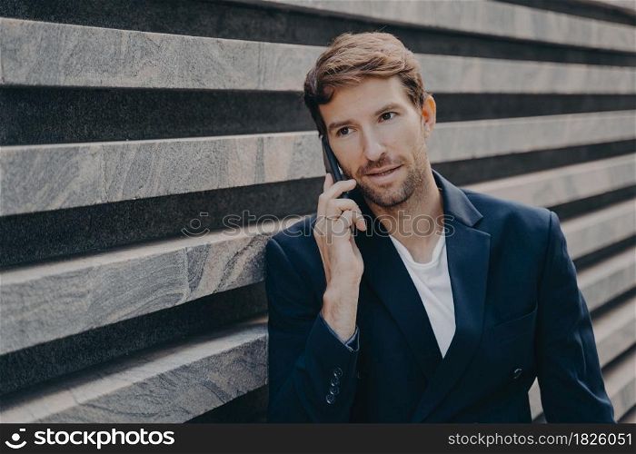 Busy professional male entrepreneur focused into distance wears formal clothes has telephone conversation stands near wall uses roaming internet connection for making international smartphone call. Busy professional entrepreneur focused into distance wears formal clothes has telephone conversation