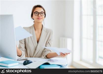 Busy successful female manager works with papers in office, poses at desktop, wears spectacles and formal outfit, busy preparing report, looks confidently at camera. Businesswoman reviews documents