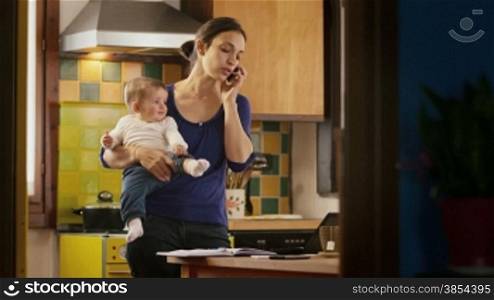 Busy woman talking on the phone in kitchen, multi-tasking mom cooking and working, holding child at home