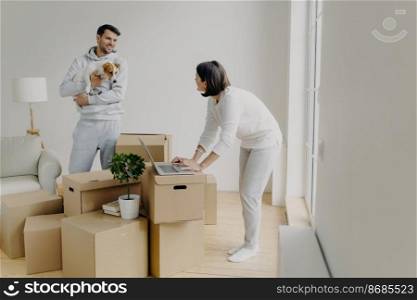 Busy woman tries to find information in laptop computer, buys furniture online, man stands with dog on hands, smiles and talks to wife, surrounded with pile of cardboard boxes have to unpack property