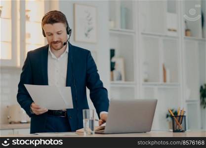 Busy young businessman in formal clothes and headset analyzing report during video chat conference, holding document while standing at his workplace at home. Bearded male employee chatting online and preparing report at remote work