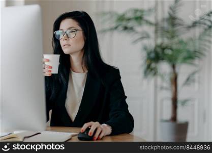 Busy young female administrative poses at desktop, drinks hot coffee from paper cup, dressed formally, develops business strategy, plans startup, involved in working process. Occupation concept