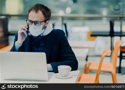 Busy young man works remotely at laptop computer during quarantine time, wears protective medical mask against coronavirus, talks via smartphone, discusses working issues, poses in empty cafe