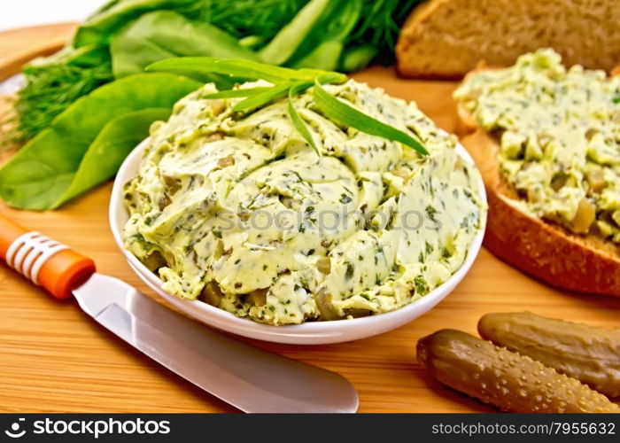 Butter with spinach, greens and pickled cucumbers in a bowl, knife, bread on a wooden boards background