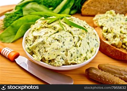 Butter with spinach, greens and pickled cucumbers in a bowl, knife, bread on a wooden boards background