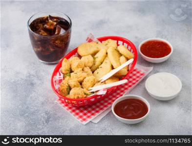 Buttered chicken nuggets and popcorn bites in red fast food basket with ketchup and glass of cola on light background with sour and cream and sweet chilli sauce.