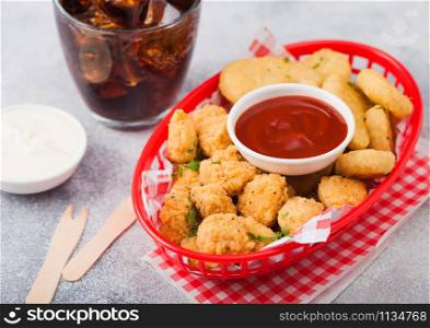 Buttered chicken nuggets and popcorn bites in red fast food basket with ketchup and glass of cola on light background with sour and cream sauce.