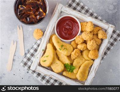 Buttered chicken nuggets and popcorn bites in white vintage wooden box with ketchup and glass of cola on light background. Fast food snack bar.