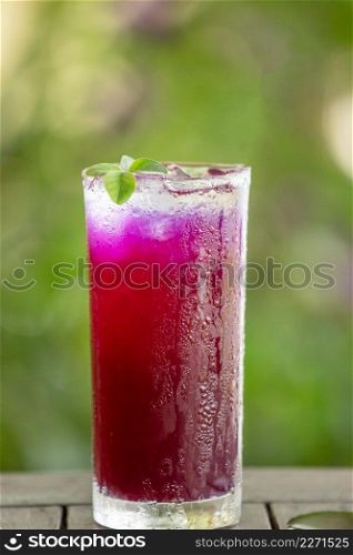 Butterfly pea juice with ice in glass, isolate on wooden table. Green leaves background. Shallow dept of field. Close. Bright sunlight.