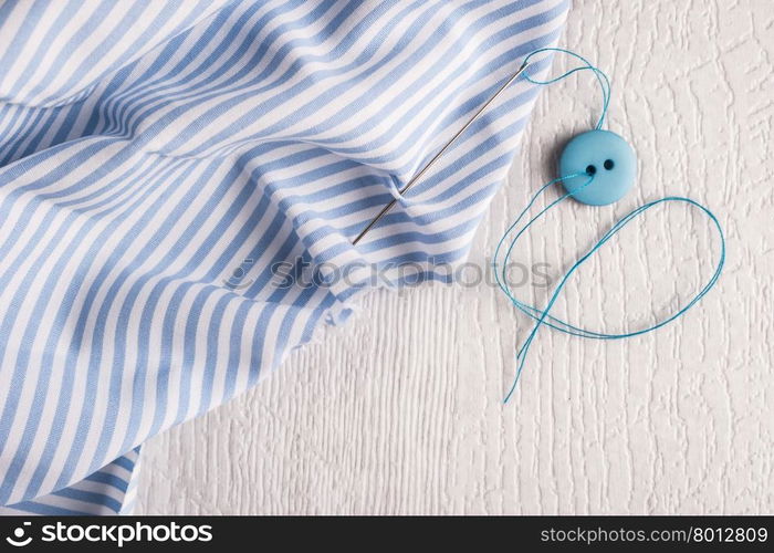 Button, thread and needle on the piece of fabric over rustic wooden table. Top view with copy space.