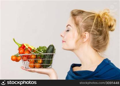 Buying good food, vegetarian products. Attractive woman holding shopping basket with green red vegetables inside, smelling with eyes closed, recommending healthy high fibre diet, on grey. Woman holds shopping basket with vegetables, smelling