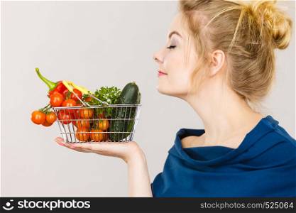 Buying good food, vegetarian products. Attractive woman holding shopping basket with green red vegetables inside, smelling with eyes closed, recommending healthy high fibre diet, on grey. Woman holds shopping basket with vegetables, smelling
