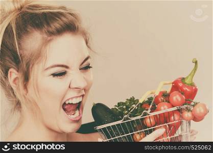 Buying good food, vegetarian products. Positive funny woman holding shopping basket with green red vegetables inside, recommending healthy high fibre diet, lifestyle modification, on grey. Woman holds shopping basket with vegetables