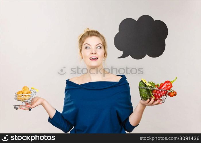 Buying healthy food, dieting products. Happy woman holding shopping cart with fruits and sweet fat bun, have to make decision, black thinking or speech bubble next to her.. Woman holding basket with fruits and sweet bun