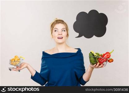 Buying healthy food, dieting products. Happy woman holding shopping cart with fruits and sweet fat bun, have to make decision, black thinking or speech bubble next to her.. Woman holding basket with fruits and sweet bun
