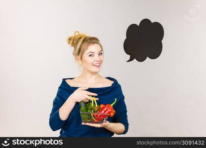 Buying healthy food, vegetarian products. Positive woman holding shopping basket with vegetables inside, standing near blank speech bubble with copy space for text, on grey. Woman holds shopping basket with vegetables, speech bubble