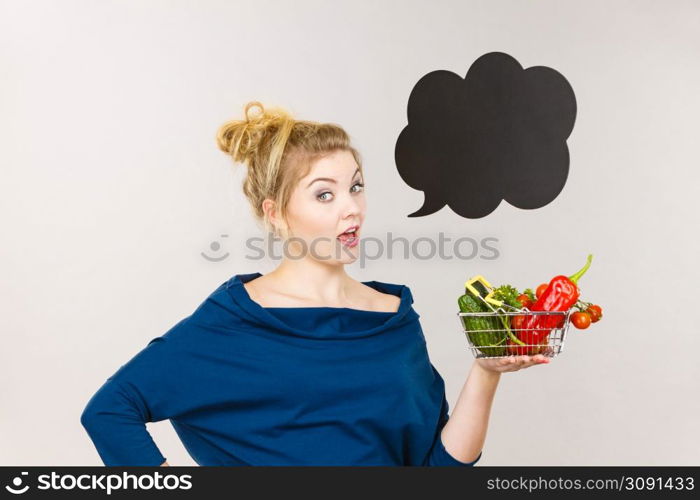 Buying healthy food, vegetarian products. Positive woman holding shopping basket with vegetables inside, standing near blank speech bubble with copy space for text, on grey. Woman holds shopping basket with vegetables, speech bubble