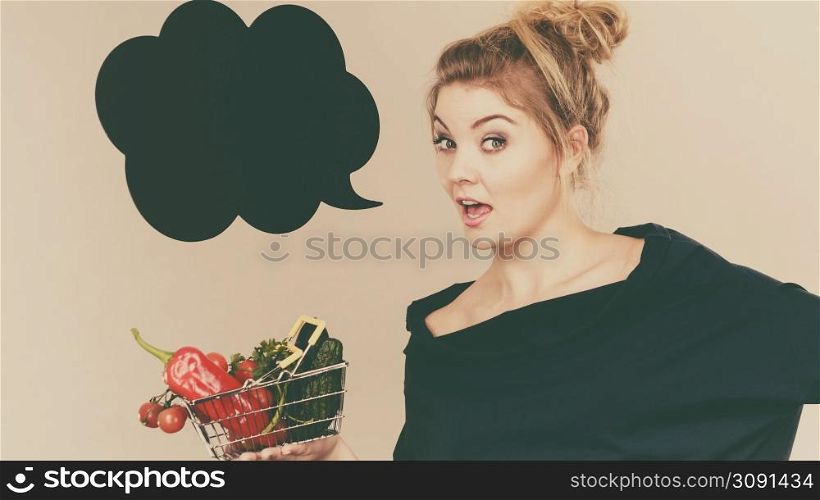 Buying healthy food, vegetarian products. Positive woman holding shopping basket with vegetables inside, standing near blank speech bubble with copy space for text, on grey. Woman holds shopping basket with vegetables, speech bubble