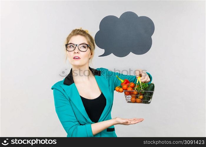 Buying healthy food, vegetarian products. Positive woman holding shopping cart with vegetables inside, standing near blank speech bubble with copy space for text, on grey. Woman holds shopping cart with vegetables, copy space