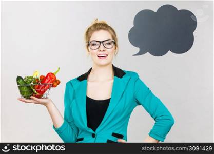Buying healthy food, vegetarian products. Positive woman holding shopping cart with vegetables inside, standing near blank speech bubble with copy space for text, on grey. Woman holds shopping cart with vegetables, copy space
