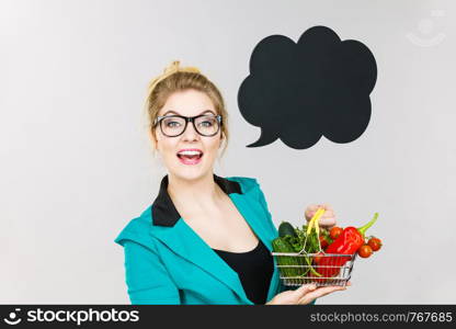 Buying healthy food, vegetarian products. Positive woman holding shopping cart with vegetables inside, standing near blank speech bubble with copy space for text, on grey. Woman holds shopping cart with vegetables, copy space