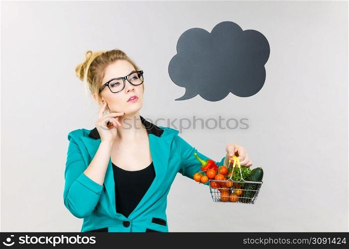 Buying healthy food, vegetarian products. Thinking woman holding shopping basket with vegetables inside, standing near blank speech bubble with copy space for text, on grey. Woman holds shopping cart with vegetables, copy space