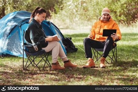 c&ing, tourism and travel concept - happy couple with tablet pc computer and book resting at tent c&. couple with tablet pc and book at tent c&