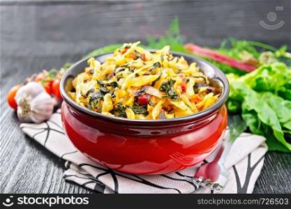Cabbage stew with leaf beets and tomatoes in a bowl on a towel, parsley, garlic and fork on black wooden board background