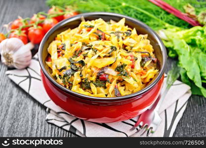 Cabbage stew with leaf beets and tomatoes in the bowl on towel, parsley, garlic and a fork on background of dark wooden boards