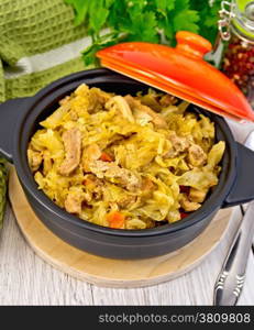 Cabbage stew with meat in a black pot with a lid, napkin, pepper, parsley. spoon on a background of light board