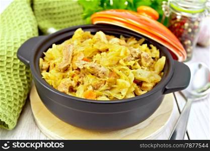 Cabbage stew with meat in black roasting pan with lid, green napkin, pepper, parsley, spoon on a light background boards