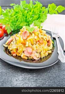 Cabbage stew with sausages in a black plate, dishcloth, tomatoes, parsley and fork on a wooden board background