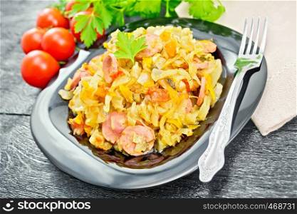 Cabbage stew with sausages in a black plate, towel, tomatoes, parsley and fork on the background of wooden boards