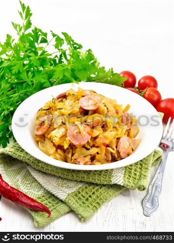 Cabbage stew with sausages in a white plate on a napkin, tomatoes, parsley and fork on light wooden board background