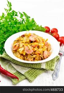 Cabbage stew with sausages in a white plate on a napkin, tomatoes, parsley and fork on light wooden board background