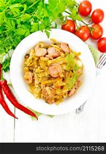 Cabbage stew with sausages in a white plate on a towel, tomatoes, parsley and a fork on the background of light wooden boards from above