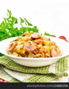 Cabbage stew with sausages in a white plate on a towel, tomatoes, parsley and a fork on the background of light wooden boards