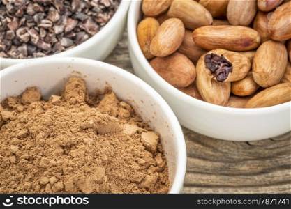cacao beans, nibs and powder in white ceramic bowls against grained wood